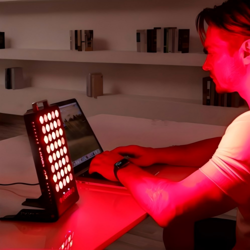 Man at desk using BestQool BQ40 portable red light therapy panel, its red LEDs glowing brightly beside his laptop.